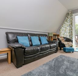 A living room with a black leather sofa with blue cushions a small black armchair with a brown throw a blue footstool wooden side tables a green lamp a bookshelf and a sliding glass door with floral curtains at Valley Lodge 5 in Callington
