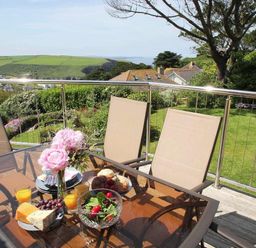 An outdoor dining table with food and drinks on a balcony overlooking a garden and green hills at Windrush in Mawgan Porth