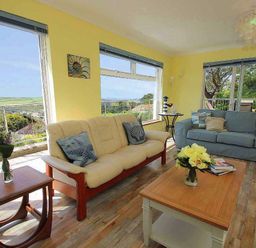 A living room with couches a coffee table with flowers and large windows showing an outdoor view at Windrush in Mawgan Porth