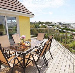 A balcony with glass table and six chairs overlooking a residential area at Windrush in Mawgan Porth