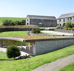 An outdoor view of a modern low building with glass walls and stone exterior surrounded by green grass and two stone houses in the background at Marhayes in Camelford