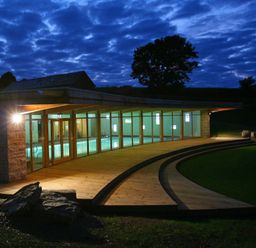 An illuminated indoor swimming pool with glass walls and wooden deck outside at Marhayes in Camelford