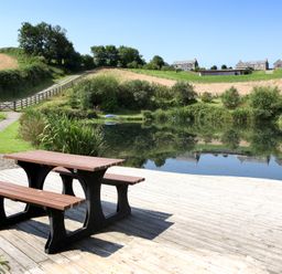 A picnic table on a wooden deck by a pond with grassy hills and buildings in the background at Westhayes in Camelford