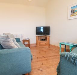 A living room with two blue sofas a wooden TV stand with a television a small wooden side table and paintings on the wall at Kerensa ha-Kres in Sennen