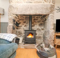 A room with a stone fireplace with a wood stove fire a blue sofa with a blanket a floor lamp a television on a wooden stand and a pet bed at Bal View in St Just