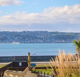 A seaside view with a playground and bench in the foreground and houses on a hillside across the water at Mackerel Sky in Marazion
