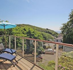 A balcony with two lounge chairs blue cushions and a blue umbrella overlooking trees and houses at Porthcurno Bay View in Porthcurno