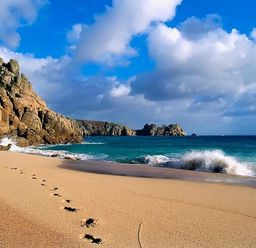 A sandy beach with footprints leading towards rocky cliffs and waves at Porthcurno Bay View in Porthcurno