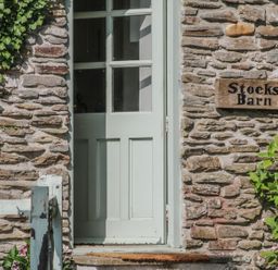 A stone wall with a pale green wooden door partly open and a sign reading Stocks Barn with green plants nearby at Stocks Barn in Looe