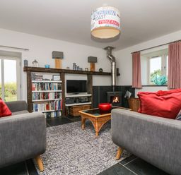 A living room with two gray sofas with red cushions a wood burning stove a bookshelf and a glass coffee table at Sundowner in Padstow