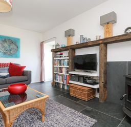 A living room with a gray sofa red cushions a wicker coffee table with a red bowl a wood burning stove and a wooden shelf with a television and books at Sundowner in Padstow