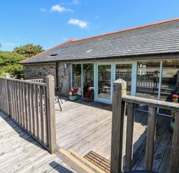 A wooden deck with railing and potted plants outside a stone building with glass doors at The Old Barn in Sennen