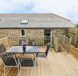 An outdoor wooden deck with a glass table and six chairs next to a stone building at Barneys Barn in Sennen