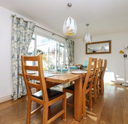 A dining room with a wooden table and chairs next to glass doors and floral curtains at Ridgeback House in Mullion
