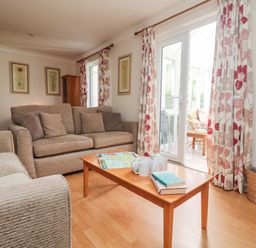 A living room with beige sofas a wooden coffee table with books and mugs curtains with floral patterns and glass doors at Tre Pol Pen in Falmouth