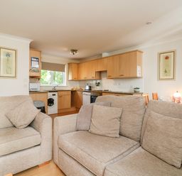 A living room with beige sofas and cushions facing a kitchen with wooden cabinets and appliances at Tre Pol Pen in Falmouth