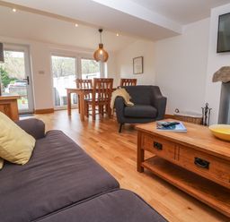 A living room with a sofa, armchair, wooden coffee table, dining table with chairs, lamp, wood-burning stove, and television at Anneth Lowen Angarrack near Hayle
