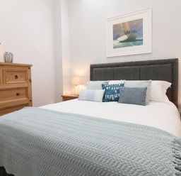 A bedroom with a bed with pillows and a blue throw blanket wooden dressers and a framed picture of a sailboat on the wall at Hazel Barn in Falmouth