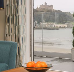 A room with a blue chair a curtain a table with a bowl of oranges and a view of a castle and water at Higher Mount View in Marazion