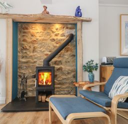 A room with a stone fireplace and wood burning stove next to a blue chair and ottoman with a patterned cushion at Higher Mount View in Marazion