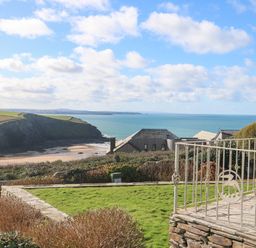 A coastal garden area with a stone patio and metal railing overlooking houses cliffs and the sea at Gwillen in Mawgan Porth