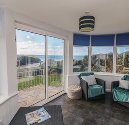 A sunroom with two armchairs and a side table by large windows overlooking a coastal landscape at Gwillen in Mawgan Porth