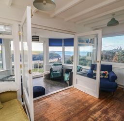A living room with armchairs and a sofa opening to a sunroom with chairs and poufs with a view of the coast at Gwillen in Mawgan Porth