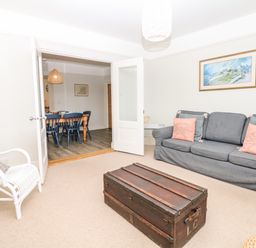 A living room with a gray sofa and coral pillows a white chair and a wooden trunk with an open door to a dining room with a wooden table and blue chairs at Cool Stone Cottage in Instow near Bideford