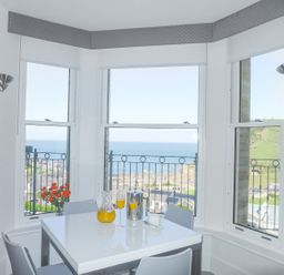 A dining area with a white table and chairs by three windows showing a sea view at 5 Arlington Villas in Ilfracombe
