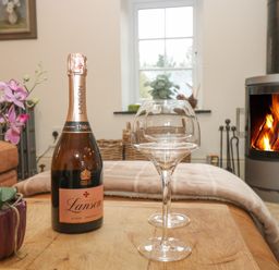 A wooden table with a bottle of Lanson champagne, two wine glasses, and a purple flower pot in a living room with a fireplace and window at Trevoole Barn in Praze An Beeble