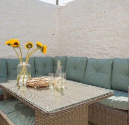 An outdoor seating area with a wicker table and corner sofa with cushions and sunflowers in a glass jar at The Crab Shell in Appledore near Bideford