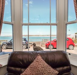A seating area with patterned curtains and a pillow overlooking parked cars and a body of water at The Crab Shell in Appledore near Bideford