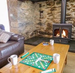 A living room with a leather sofa a wooden coffee table with a scrabble game and mugs and a wood burning stove set in a stone wall at Tai Ucha in Cerrigydrudion