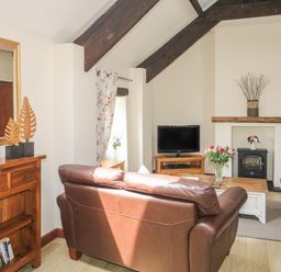 A living room with a brown leather sofa a wooden cabinet a television and flowers on a table at The Hayloft in St Neot near Dobwalls