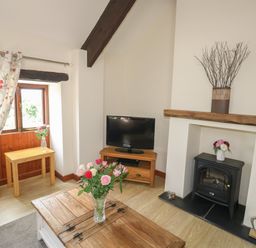 A living room with a wooden coffee table with flowers on it a small wooden table by a window a television on a stand and a fireplace with decorations at The Hayloft in St Neot near Dobwalls