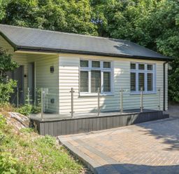 A small house with cream siding windows and a glass railing on a deck near a paved driveway and green trees at Hidden Gem Cabin in Lettermacaward near Dungloe County Donegal