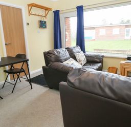 A living room with two black sofas a small dining table with two chairs and a window with blue curtains at 21 Siesta Mar in Mundesley
