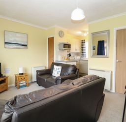 A living room with brown leather sofas a television on a wooden stand a small wooden side table and a kitchen area in the background at 21 Siesta Mar in Mundesley