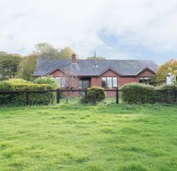 A single-story brick house with a slate roof behind a black fence and bushes at The Alders in Llanfyllin
