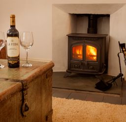 A room with a wood burning stove firewood bucket fireplace tools and a table with a wine bottle and two glasses at Star House in Marazion