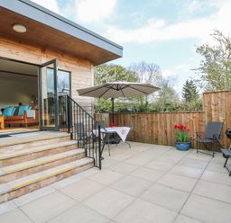 A patio with chairs a table an umbrella and plants outside a wooden house at No1 The Lodge in Acton near Bishops Castle