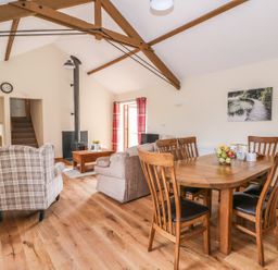 A dining area with a wooden table and six chairs next to a living room with sofas a plaid armchair a wood stove and exposed wooden beams at Pentre Fach in Llandrindod Wells