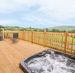 An outdoor wooden deck with a hot tub a lounge chair and a table with chairs and umbrella overlooking green hills at Pentre Fawr in Llandrindod Wells
