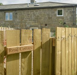 A wooden fence gate with a sign reading Rose Cottage and a stone building in the background at Rose Cottage in Edlingham