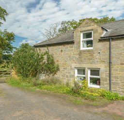 Exterior of a stone house with white-framed windows and a slate roof next to a dirt driveway and wooden gate surrounded by trees at Rose Cottage in Edlingham