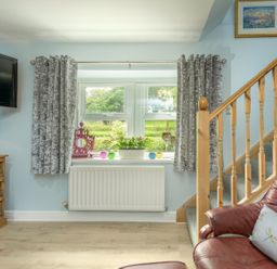 A living room window with grey curtains a wooden staircase a brown leather chair a wooden cabinet with a lamp and decorative items at Rose Cottage in Edlingham