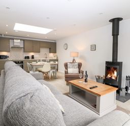 An open-plan living room and kitchen with a grey sofa a wood stove a dining table and kitchen cabinets at Rabbit's Warren in Titchfield Common