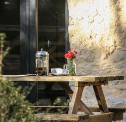 A wooden picnic table with a glass coffee press a white cup and saucer and a jar of red flowers outside by a stone wall