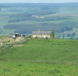 A stone cottage on a green hill with vehicles and outbuildings surrounded by fields and distant hills at Humbleton Cottage in Haydon Bridge near Hexham