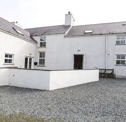 An exterior view of a white house with multiple windows a chimney two skylights a gravel yard and a low white wall at Bodegri Cottage in Llanrhyddlad near Church Bay